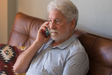 Serious senior aged man doing phone call sitting at home with smart phone - sad emotion elderly people lonely indoor - caucasian old male use modern device sitting on the sofa