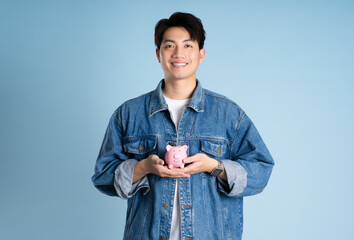 Portrait of an Asian guy holding a piggy bank and posing on a blue background