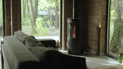 Cozy house chimney fire place with fire burning at the living room in tiny house, wooden wall and large window on the background, view to the green forest at summer, slow motion.