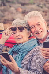 Cheerful old senior couple use phones together outdoor with red car in background - concept of travel mature people and modern technology in elderly retired happy lifestyle