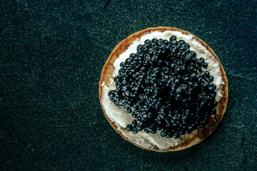 A blini with caviar and cream cheese, overhead flat lay shot on a black slate background with a place for text