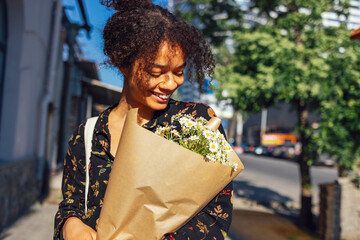 Young smiling afro american woman walks along the street with a bouquet of daisies.