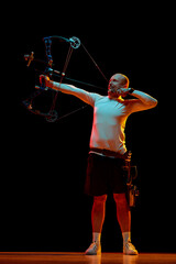 Full-length of man, archer in motion, training, aiming archery bow on target against black studio...