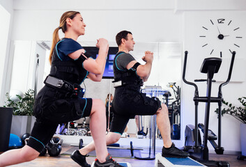 Fototapeta na wymiar Sport couple in ems suits is engaged in physical activity in fitness club or gym. Man and woman with clenched hands doing forward lunges with step platforms.