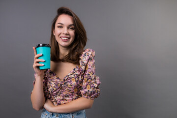 An attractive young brunette woman with beautifully styled hair and a stylish shirt holds a paper cup for coffee. Gray background indoors