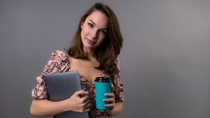 A young woman with a laptop on a grey background. The model drinks coffee from a paper cup. Copy Space