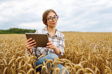 Naklejka na ściany i meble A young female agronomist with a modern tablet checks the quality and growth of the crop in a wheat field. Gardening concept, harvesting.