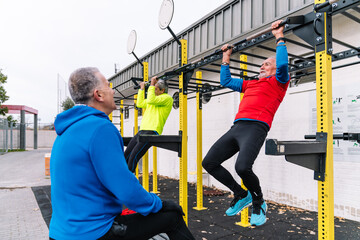 Three senior men exercising at an outdoor fitness station, with one actively doing pull-ups.