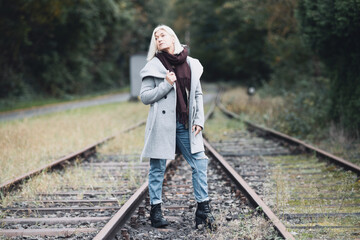Naklejka na ściany i meble Older woman in her fifties with blonde hair, dressed in a long gray coat and black scarf, stands between railway tracks, full body portraits.
