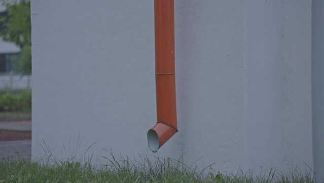 Metal pipe for rainwater drain runs along grey concrete wall. Buildings covered with red bitumen roofing sheets locate nearby closeup