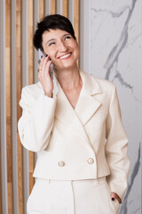 Business woman is standing in her office in a white pantsuit talking on a mobile phone and smiling.