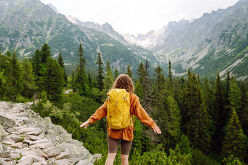 Naklejka premium Happy woman with a yellow hiking backpack enjoying the mountain landscape. A young traveler travels along mountain paths. Adventure, travel concept.