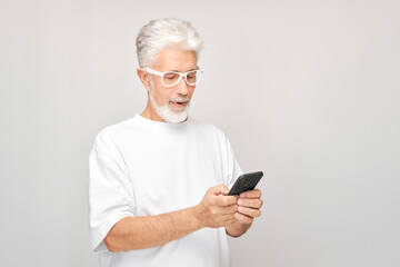 Fototapeta premium Portrait of gray-haired man in white T-shirt looks on mobile phone and thinks. Person with smartphone isolated on white background.