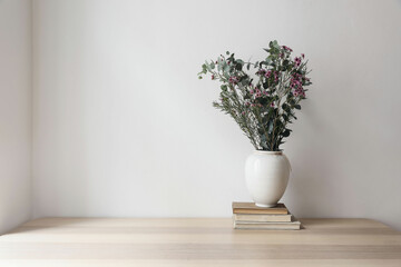 White vase with bouquet of eucalyptus tree brnahces and waxflowers. Old books on wooden table. Empty beige wall background. Neutral mockup. Scandinavian home, living room design. - obrazy, fototapety, plakaty