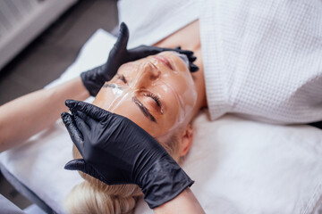 Close up cosmetologist hands in black gloves apply transparent vitamin mask to her client face in spa salon or clinic.