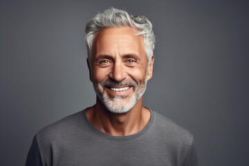 Handsome mature man with grey hair and beard smiling at camera while standing against grey background - obrazy, fototapety, plakaty
