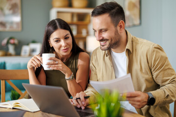 Married couple sit at a table in the living room with laptop,working from home office - 705025304
