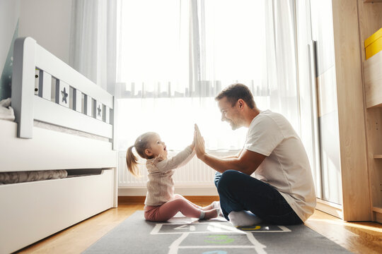 Supportive dad and his little girl are sitting on the floor in baby room and giving high five to each other. - Powered by Adobe