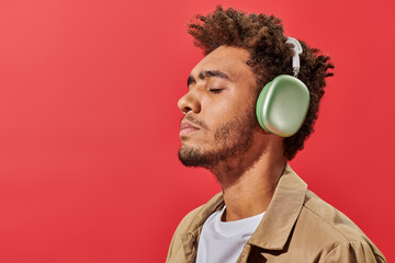 portrait of young african american man in wireless headphones listening music on red background