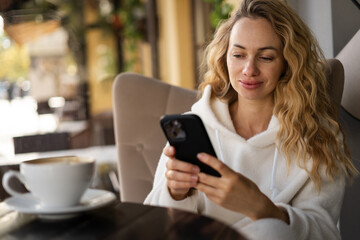Beautiful blonde woman holding cell telephone typing text message or promotional content, lady watching video on mobile phone during coffee break in cafe or coffee shop