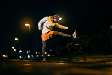 An urban night runner is stretching his leg outside.