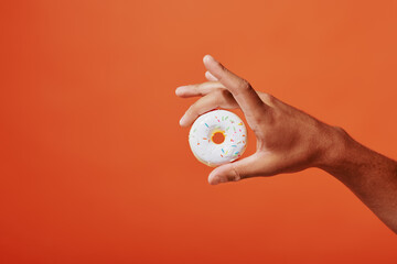 cropped shot of person holding glazed vanilla donut with sprinkles on orange background, white icing