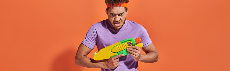 african american man in headband playing water fight with toy gun on orange background, banner