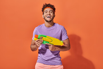 happy african american man in headband playing water fight with toy gun on orange background