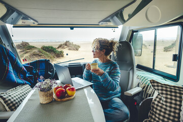 Living and working inside your camper while traveling and a digital nomad-free lifestyle. A woman sitting in an RV enjoys relaxing and connecting to her laptop. The beach in the background outside - obrazy, fototapety, plakaty