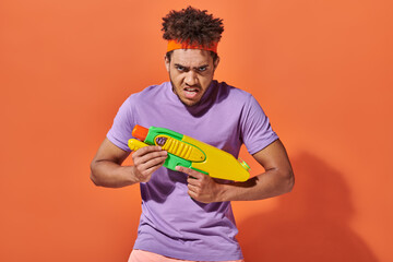 african american man in headband playing water fight with toy gun on orange background, grimace