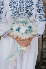 A woman wearing an embroidered dress holds a bouquet. Wedding bouquet flowers on background stylish bride. Wedding ceremony outdoors. Closeup.