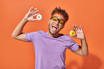 happy young african american man in sunglasses holding small tasty donuts on orange background