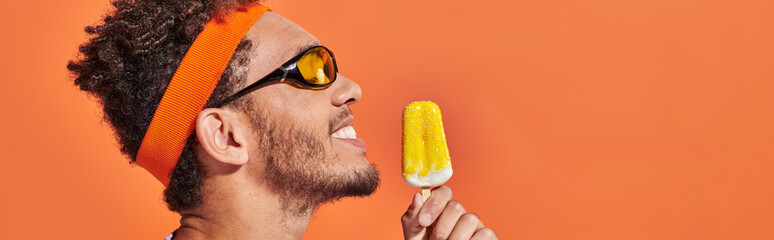 happy african american man in sunglasses holding frozen ice cream on orange backdrop, banner