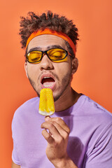 african american man in sunglasses and headband eating fruity ice cream in summer on orange backdrop
