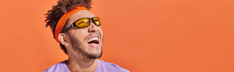 african american man in sunglasses and headband laughing and looking away on orange backdrop, banner