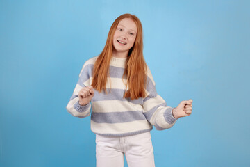 Fototapeta premium Smiling redhead teenage girl dancing on blue background in studio.