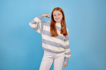 Smiling redhead teenage girl raises arms and shows biceps on blue background in studio.