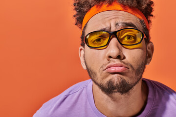 upset african american man in eyeglasses and headband grinning on orange background, grimace