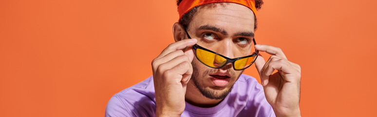 annoyed african american man in eyeglasses and headband rolling eyes on orange background, banner - obrazy, fototapety, plakaty