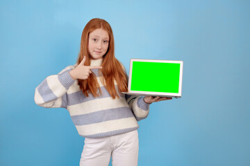 Smiling redhead teenage girl holding laptop on blue background in studio. Green screen on laptop.