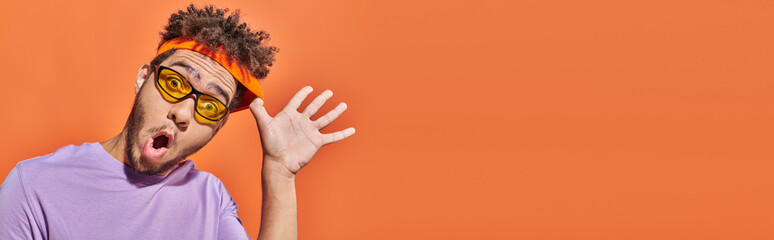 banner, african american man with surprised face expression adjusting headband on orange background