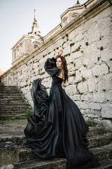 Female in long black dress stands on stairs near ancient palace at sunset. Model girl in nature autumn day. Luxury woman near old Pidhirtsi Castle, Lviv region, Ukraine. Stylish bride. Back view