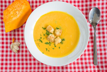 Top view for white bowl with pumpkin soup, slice of pumpkin on a checkered tablecloth
