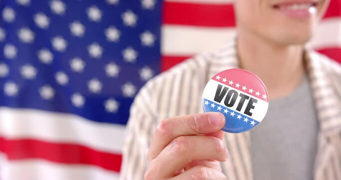 Happy biracial man in front of american flag holding badge with vote text, copy space, slow motion