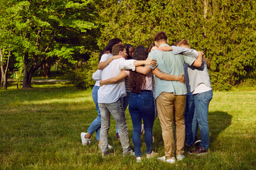 Fototapeta premium Team of young diverse people huddling in park all together. Group of friends standing and hugging on fresh green lawn in beautiful summer park. Teamwork, support, friendship concept