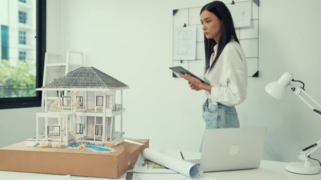 Beautiful caucasian architect focus on using tablet to check house model on meeting table with blueprint, house model and architectural document scatter around at meeting room. Immaculate.