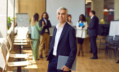 Portrait of happy smiling confident mature business man looking at the camera holding a folder with financial documents with a team of company employees talking in background in office.