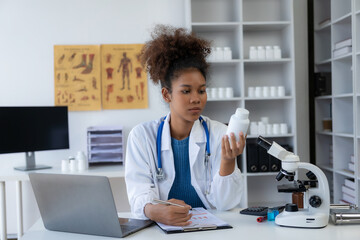 A young African American female doctor in a white coat is sitting at a consulting table using a...