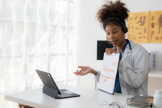 A young African American female doctor in a white coat is sitting at a consulting table using a laptop. Telemedicine service Doctor chats with patient via online video conference