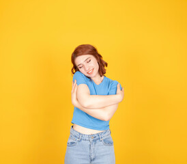 Hugging herself, close up portrait of young attractive cheerful woman enjoying hugging herself. Lovely charming red bob haired girl wear blue t-shirt, jeans. Isolated yellow background.